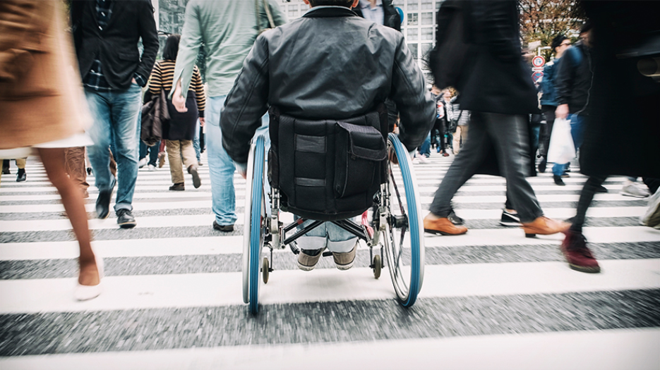 A person in a wheelchair crossing a busy street intersection while pedestrians walk in multiple directions around them.
